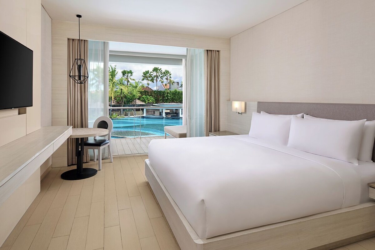 Une chambre d’hôtel avec un grand lit, un téléviseur fixé au mur, une petite table avec une chaise, et une vue sur une piscine et la verdure environnante.