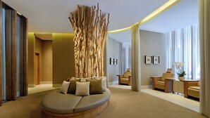 Ein modernes Hotelzimmer mit einer gebogenen Sitzecke, einem Holzkunstobjekt, einer Couch mit Kissen, einem Beistelltisch, einem Sessel und Blick ins Freie durch große Fenster.