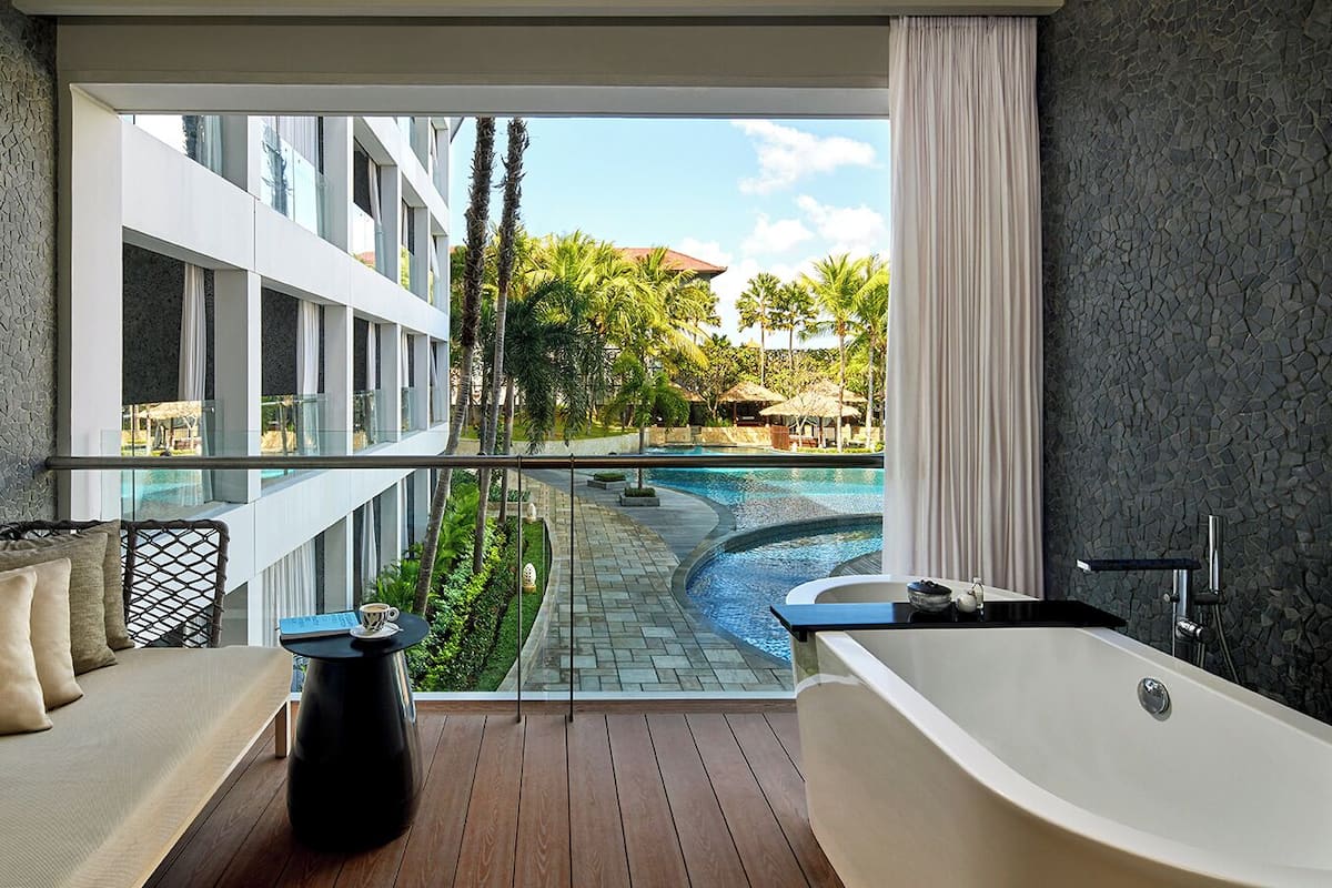 Une salle de bain moderne avec une baignoire, un coin salon avec des coussins et une vue sur une piscine et un bâtiment.