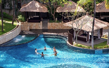 A swimming pool with a stone border, palm trees, and thatched-roof structures.