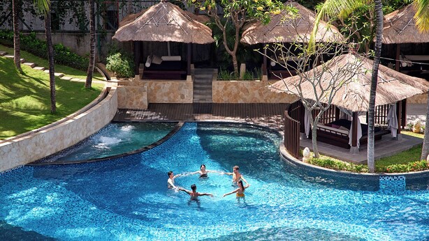 Ein Pool mit Steinrand, Palmen und strohgedeckten Gebäuden.