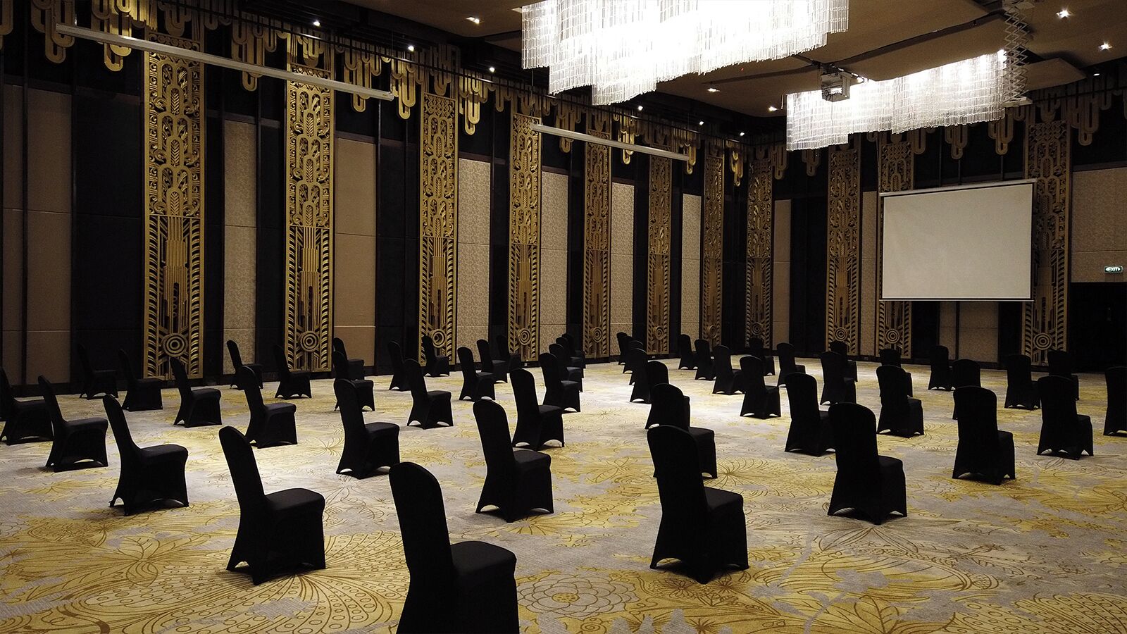 Un salón de conferencias con filas de sillas negras, una pantalla grande y decoraciones ornamentales en las paredes.