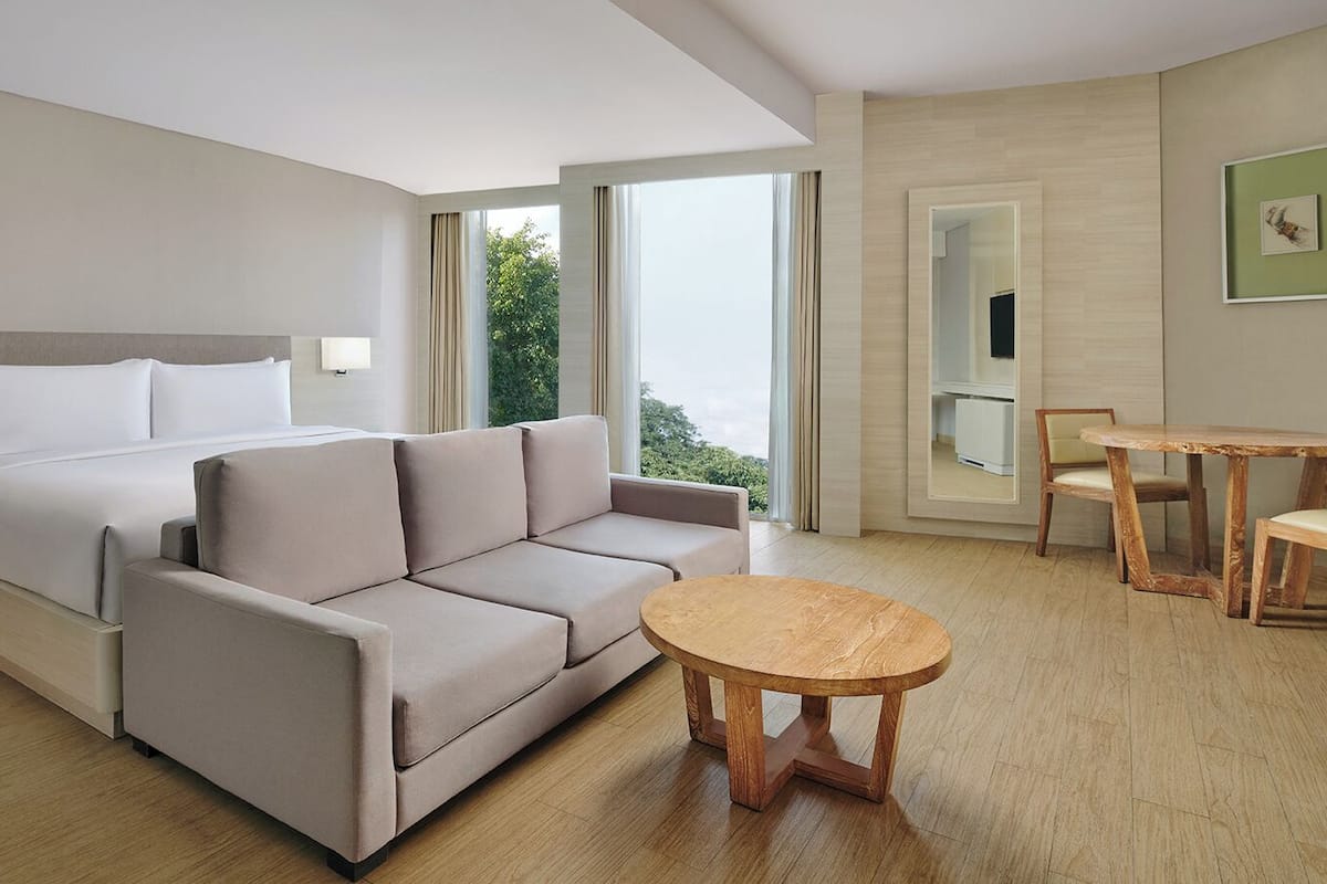 Une chambre d’hôtel moderne, équipée d’un lit, d’un canapé, d’une table ronde en bois et offrant une vue sur la verdure par la fenêtre.