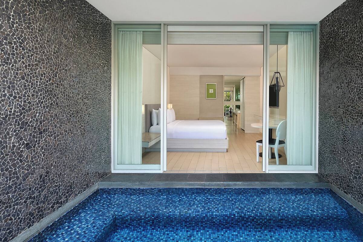 Une chambre d’hôtel moderne dotée d’une grande porte-fenêtre coulissante donnant accès à une chambre, à un petit coin salon et à une zone piscine.