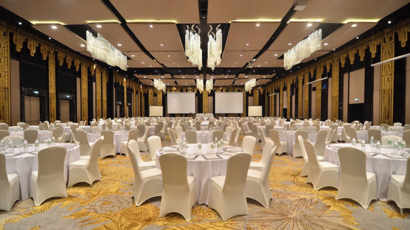 Un amplio salón de banquetes con mesas redondas y sillas blancas, suelo alfombrado y candelabros.