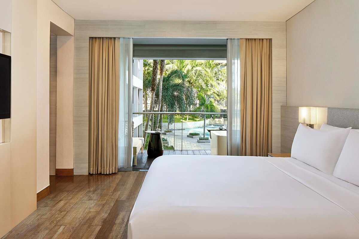 Une chambre d’hôtel avec un grand lit, un téléviseur fixé au mur et une vue sur une piscine et des palmiers à l’extérieur.