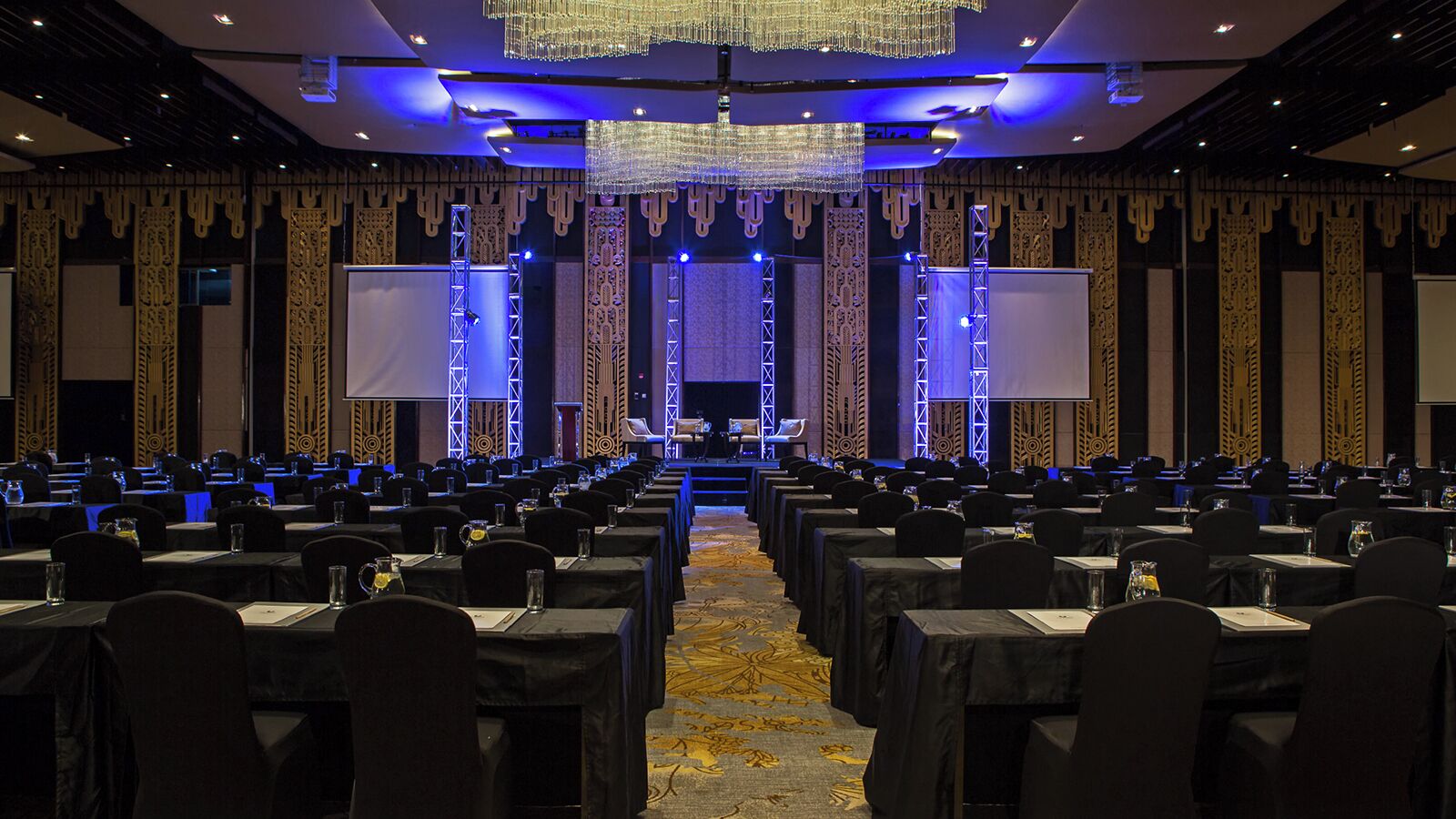 A conference room with rows of tables and chairs, a stage with a screen, and decorative wall panels.