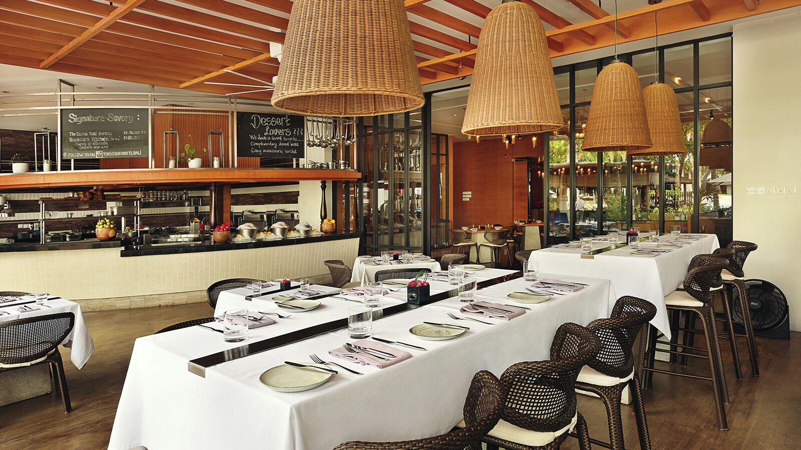 Un restaurant avec de longues tables dressées pour le repas, des chaises en osier et des lampes en osier suspendues au plafond.