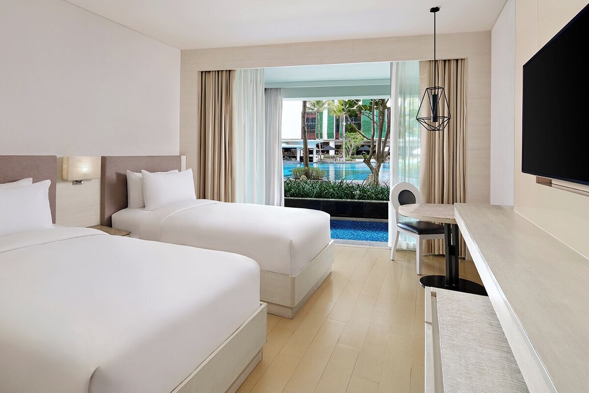 Une chambre d’hôtel avec deux lits, un bureau, une télévision et une vue sur l’espace extérieur de la piscine.