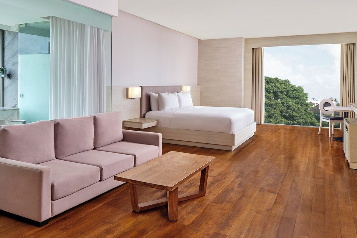 Une chambre d’hôtel moderne avec un plancher en bois, un canapé, un lit, un bureau et une grande fenêtre donnant sur la ville.