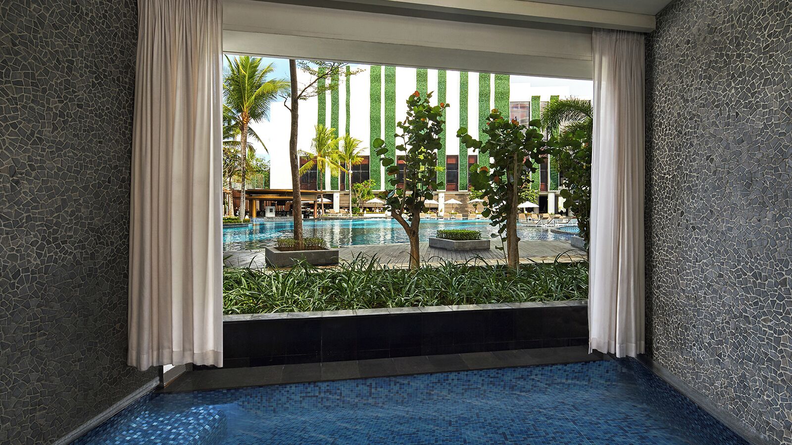 A room with a view of an outdoor swimming pool, a balcony with a view of the pool, and a window with curtains.