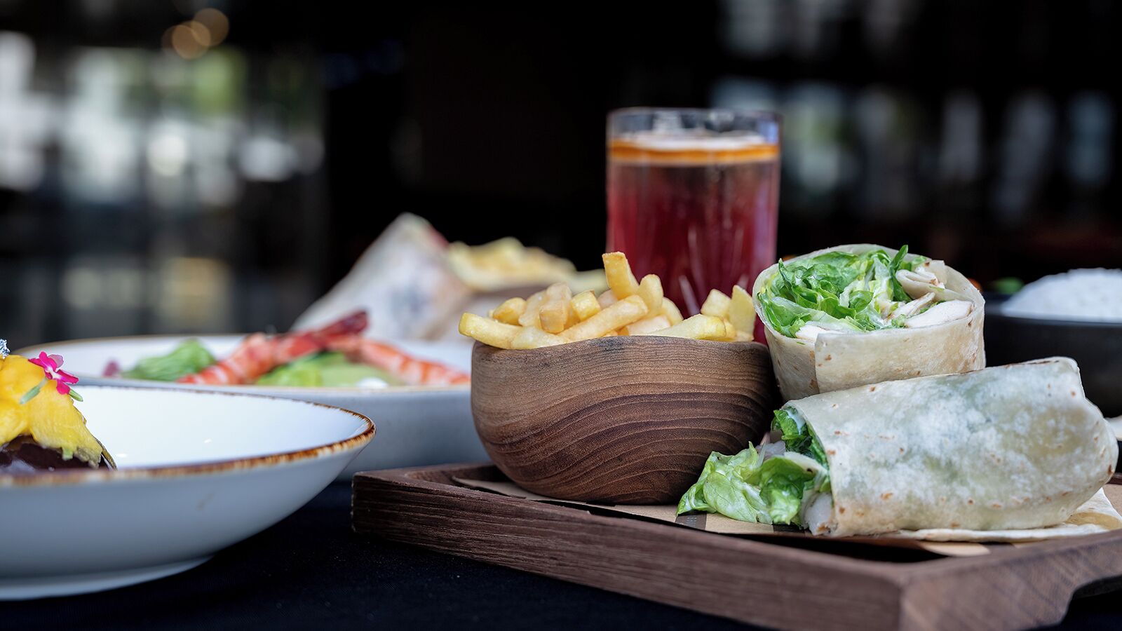 Una mesa con comida, incluyendo un tazón de papas fritas, un vaso de bebida roja y wraps.