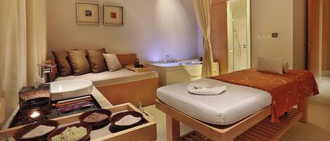 A hotel room with a bed, a sofa, a small table with bowls, a sink, and a shower area.