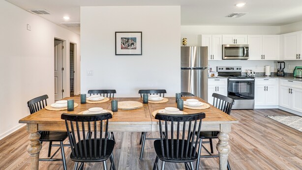 Un coin repas avec une table en bois et des chaises noires, une cuisine avec des meubles blancs et des appareils en acier inoxydable, et un tableau encadré accroché au mur.