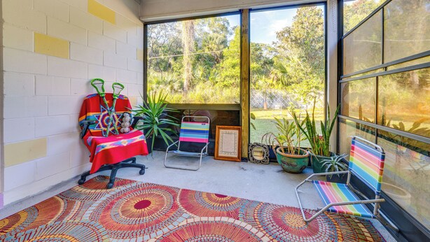 A sunlit room with a patterned rug, a table with a colorful tablecloth, two chairs, and potted plants.