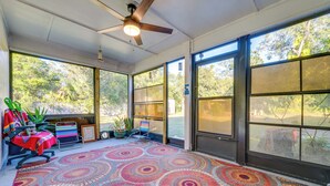 A sunlit room with large windows, a ceiling fan, a colorful rug, and a chair with a red tablecloth.