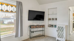 Una habitación con una televisión instalada en la pared, una mesa consola metálica con cajones, un armario blanco con puertas de vidrio y una ventana con cortinas.