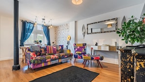 A living room with a colorful sofa, a black rug, wooden flooring, a dining area with a table, chairs, and a potted plant. - Pontypool Haven Retreat - 3Bedroom Flat (Torfaen Principal Area)