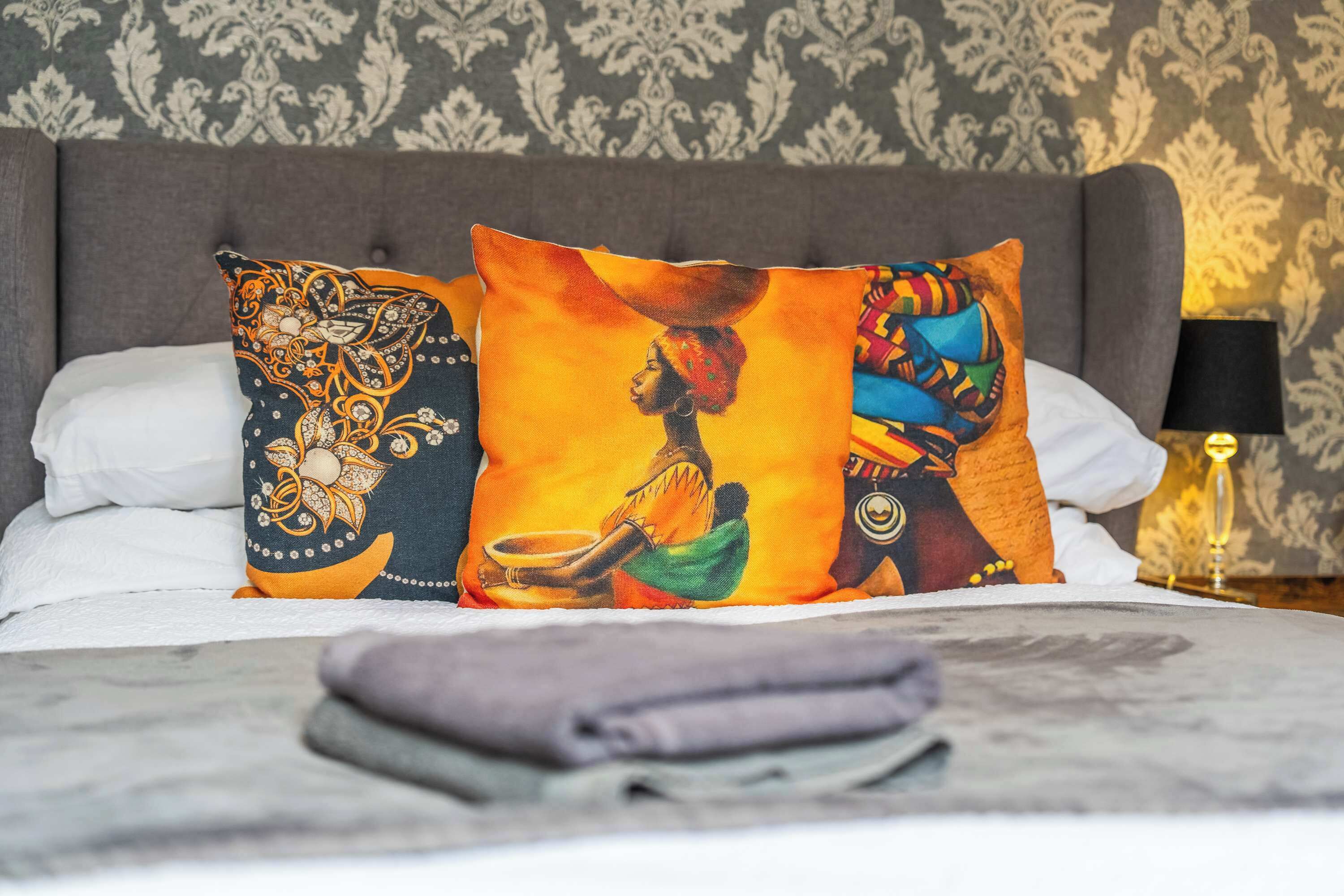 Una cama con almohadones decorativos, una lámpara y un papel tapiz estampado.