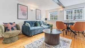 Un salon moderne avec un canapé bleu, une table basse ronde et des fauteuils orange. Des œuvres d’art encadrées ornent le mur, et une fenêtre offre une vue sur les arbres.