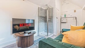 Un salon moderne doté d’un téléviseur à écran plat, d’une cabine de douche en verre et d’un canapé vert agrémenté de coussins jaunes.