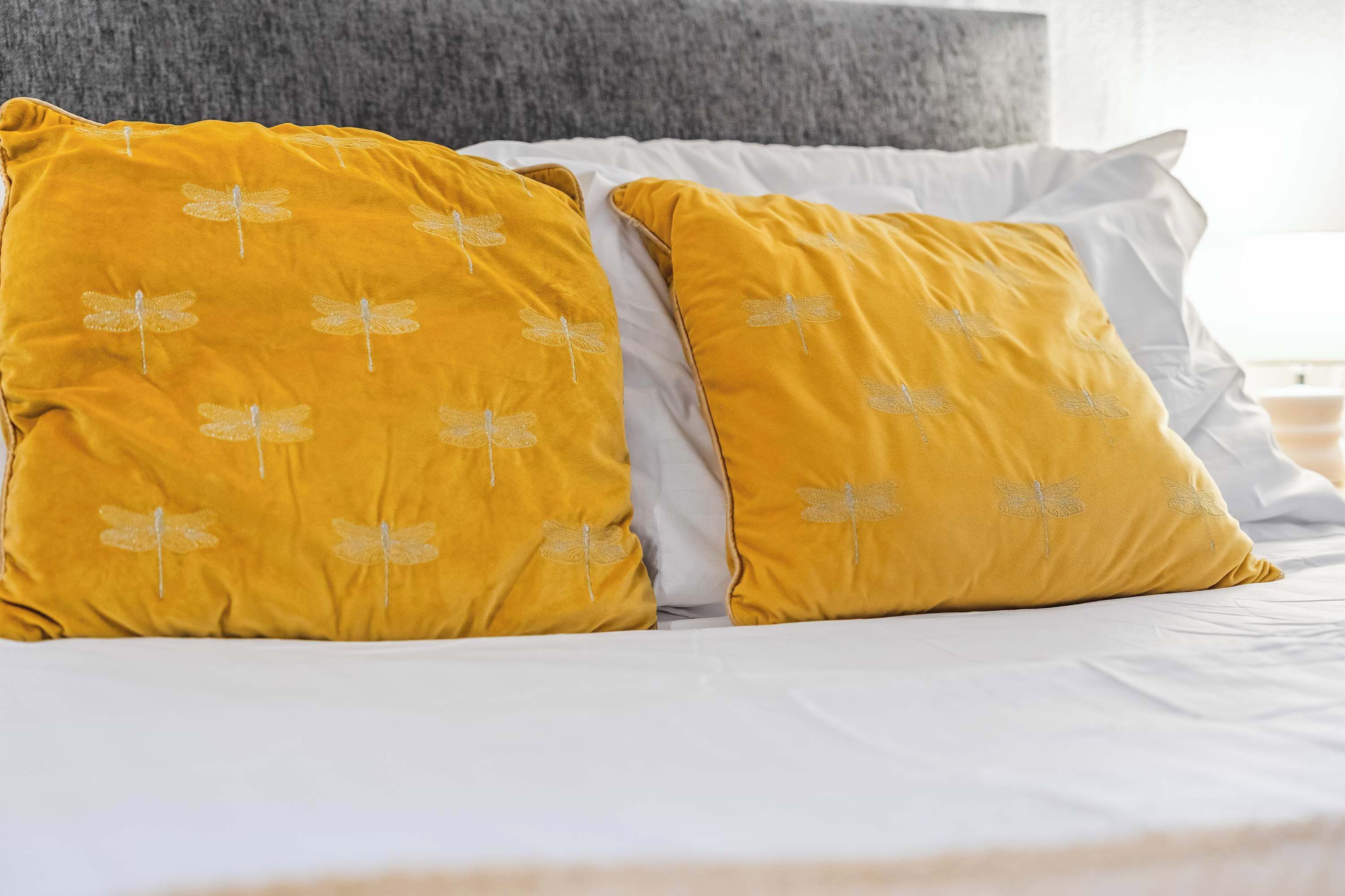Een bed met wit beddengoed en twee mosterdgele kussens met libellenmotieven.