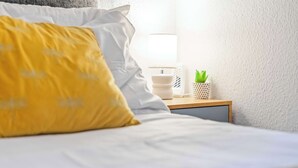 Una cama bien hecha con almohadas blancas y amarillas, una mesita de noche con una lámpara, una planta pequeña y una pared blanca.