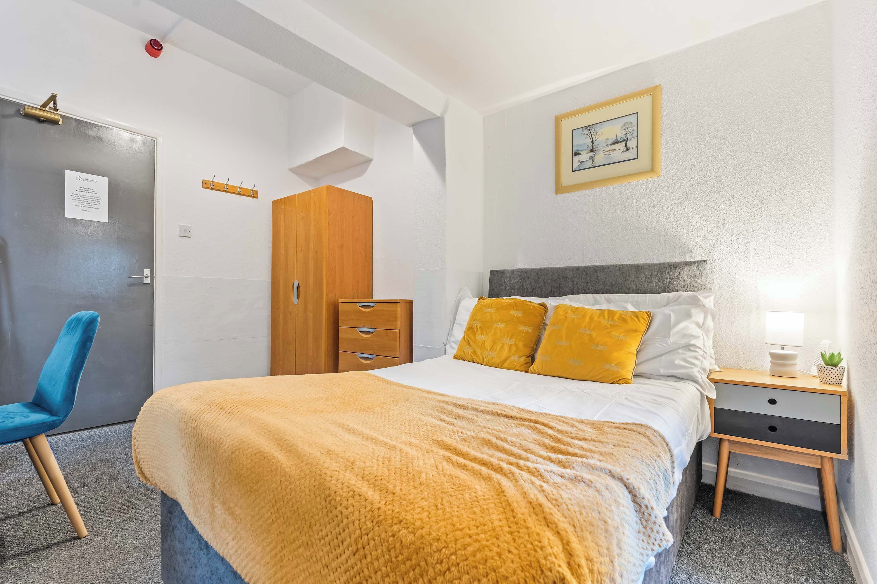 A bedroom with a bed, a nightstand, a wooden wardrobe, a blue chair, and a door with a sign.