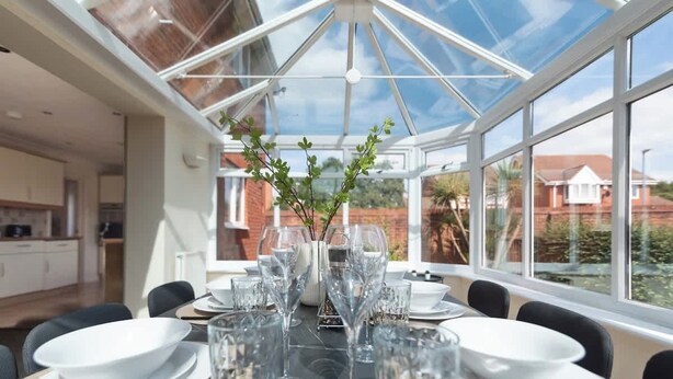 Un espace repas doté d’un plafond en verre, une table dressée pour six personnes avec de l’argenterie blanche et des verres, et un vase contenant des feuilles vertes.