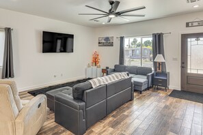 A modern living room with a gray sectional sofa, a beige armchair, a flat-screen TV mounted on the wall, a ceiling fan, and a large window with curtains.