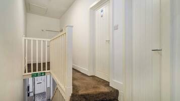 A narrow hallway with white walls, a carpeted staircase, and a fire exit sign.