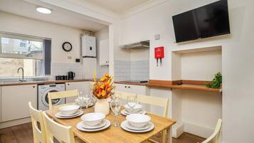 A modern kitchen with white cabinets, a built-in washing machine, a TV mounted on the wall, and a dining area set with white plates and glasses.