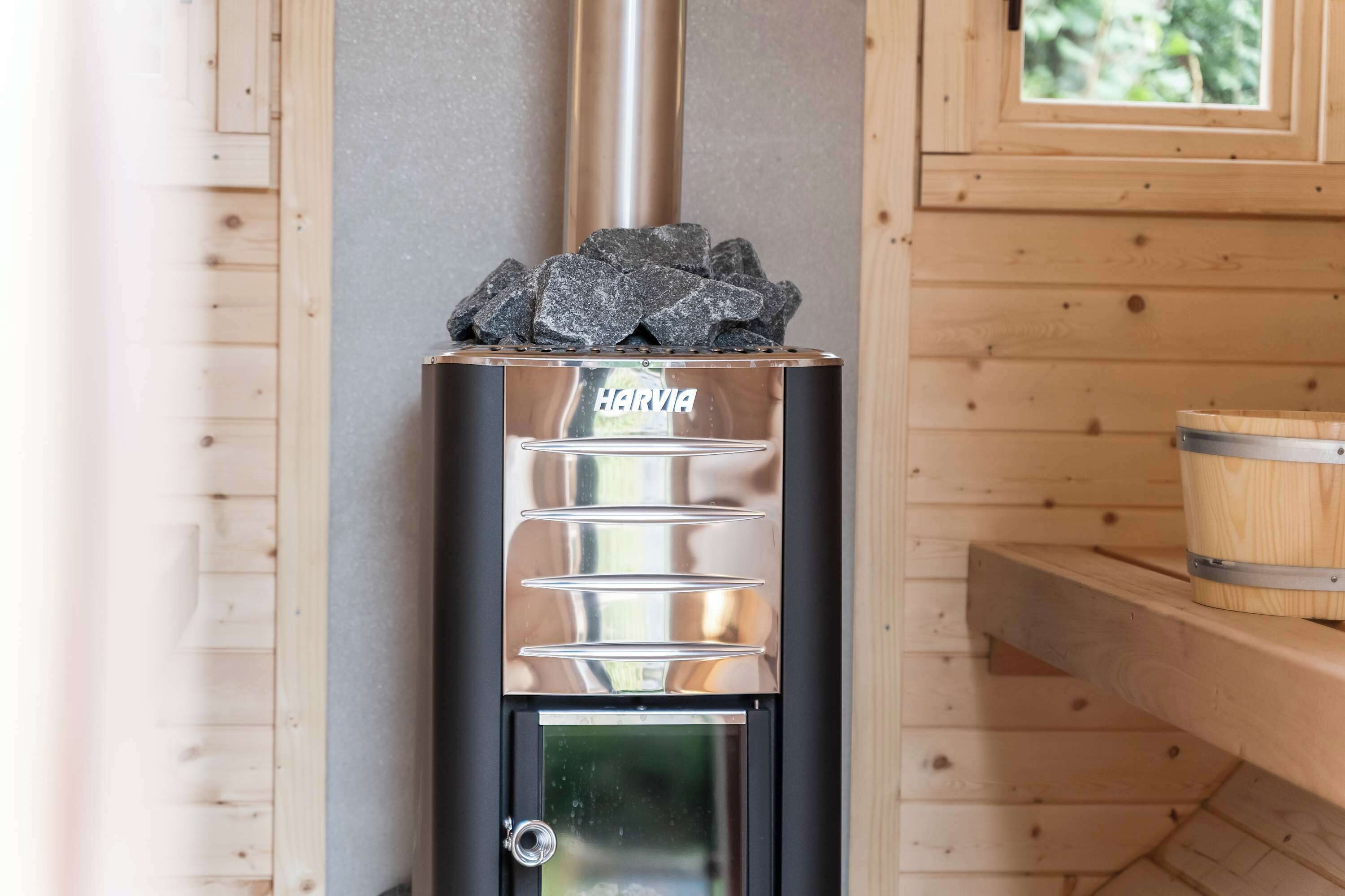 Een Harvia houtkachel met een metalen schoorsteen, een glazen deur en een houten plank met een vat.