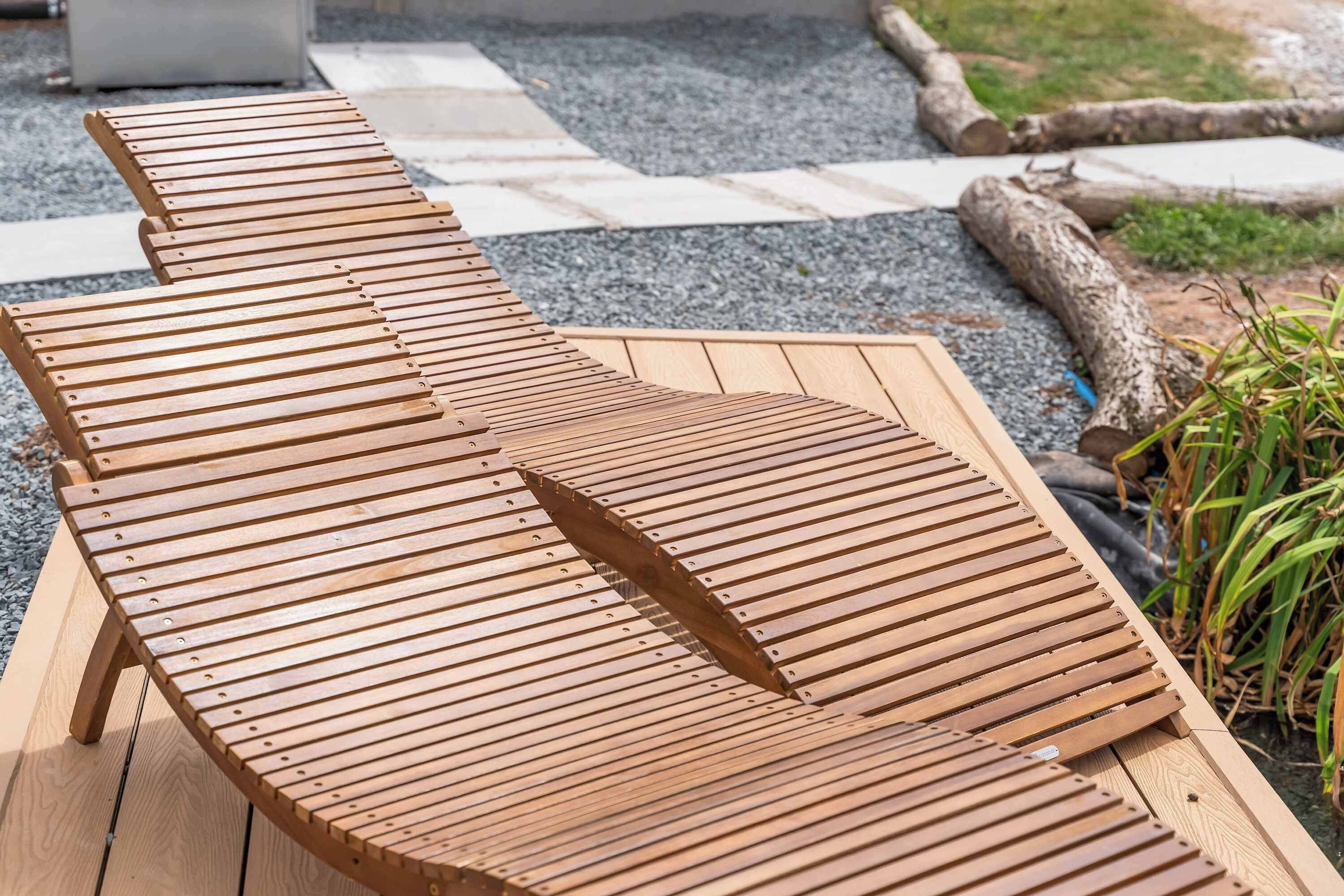 Un fauteuil de jardin en bois, doté d’une assise à lattes, placé sur un sol de gravier, avec un chemin de pierres en arrière-plan.