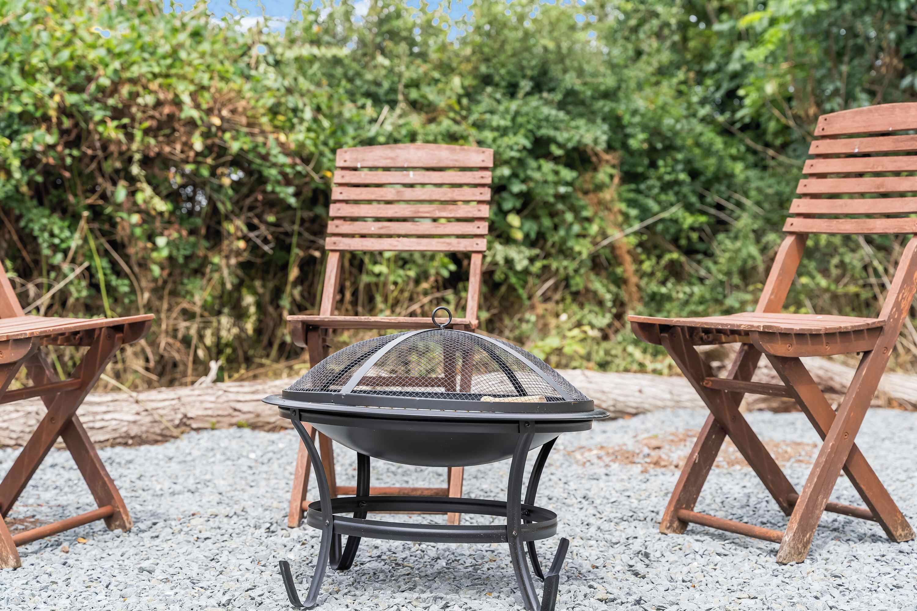 A grill with a metal grill cover, surrounded by three wooden chairs on a gravel surface.