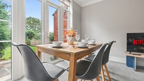 A dining area with a wooden table set for four, a television displaying 'NETFLIX', and a view of a brick house through large glass doors. - 4-Bed Beeston Getaway | Free Parking (Beeston)