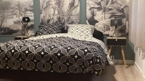 Une chambre à coucher avec une couvre-lit à motifs, une fresque murale et un petit bureau avec une lampe.