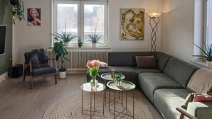 Una sala de estar con un sofá gris, una mesa de centro redonda y una silla. Hay dos plantas en macetas sobre el alféizar de la ventana y una lámpara de pie.