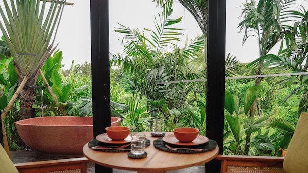 A tropical setting with a round wooden table, two chairs, a large planter, and a view of lush greenery through a glass door. - Patha (Canggu)