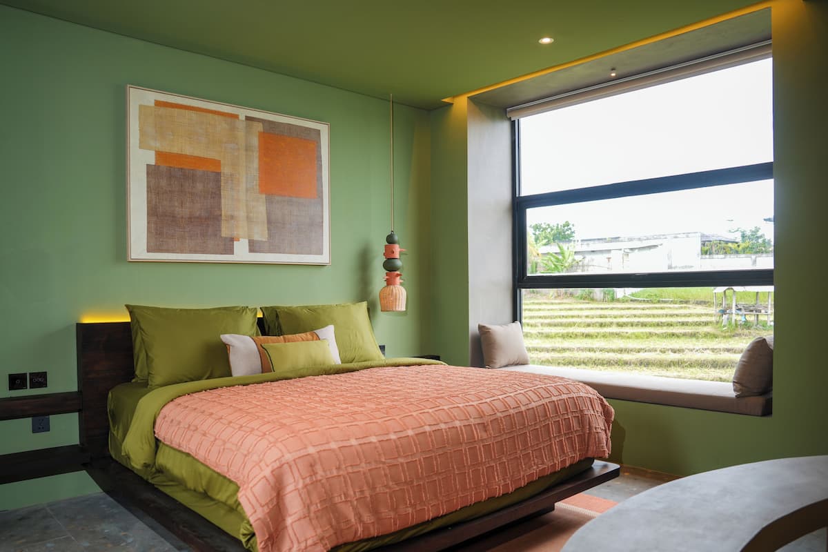 Une chambre moderne avec un grand lit, des murs verts, une fenêtre donnant sur un jardin et un tableau abstrait encadré au-dessus du lit.