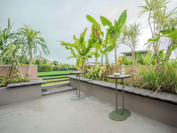 An outdoor patio with two round tables, surrounded by various tropical plants and a view of a green landscape. - Patha (Canggu)
