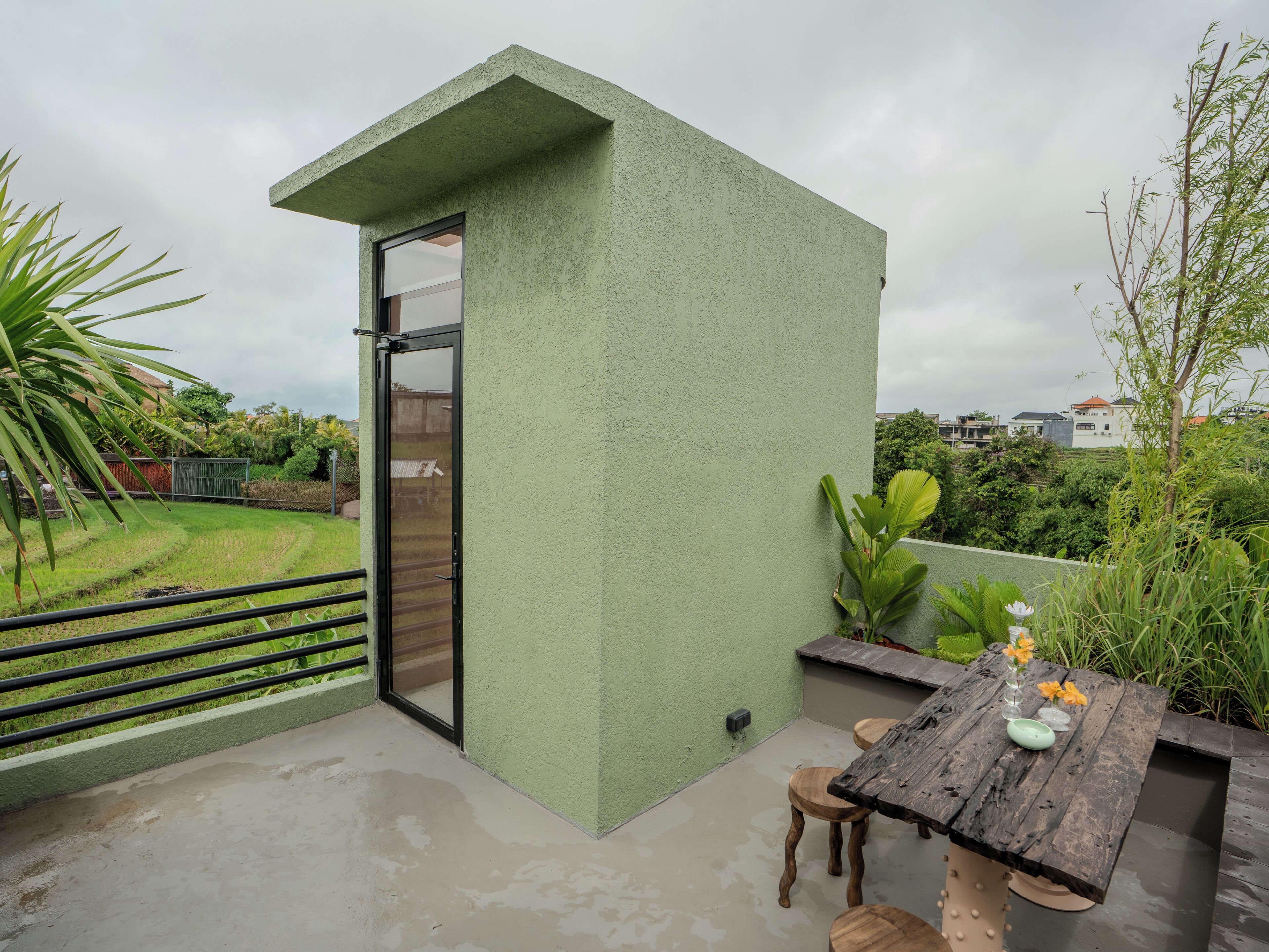 A balcony with a green textured wall, a sliding glass door, a wooden table with chairs, and a small garden area with plants and a wooden fence.