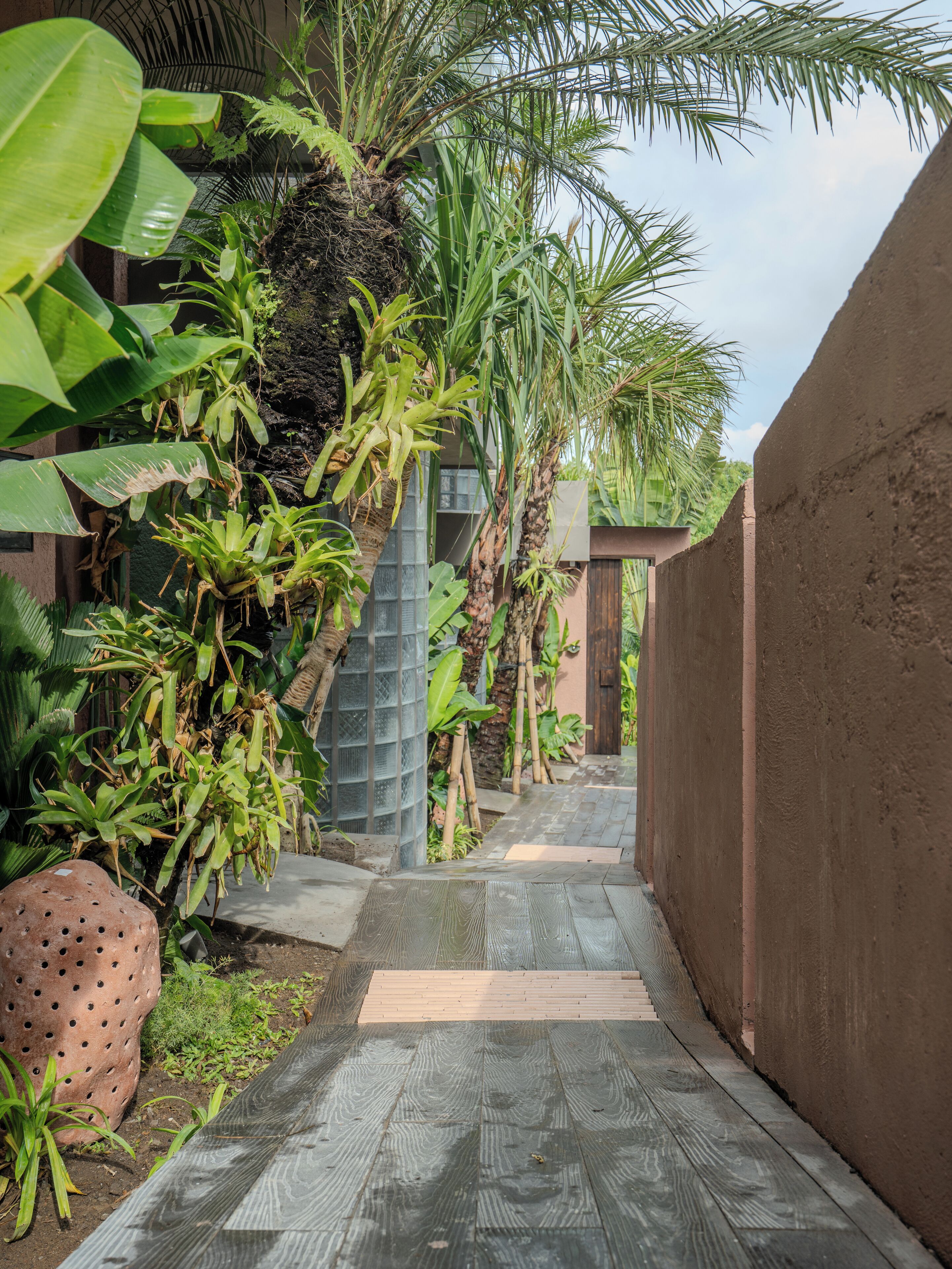 A narrow pathway flanked by lush greenery and a concrete wall, leading to a building with a metal fence.