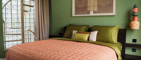 A bedroom with a bed, a window with curtains, a painting on the wall, and a hanging lamp.