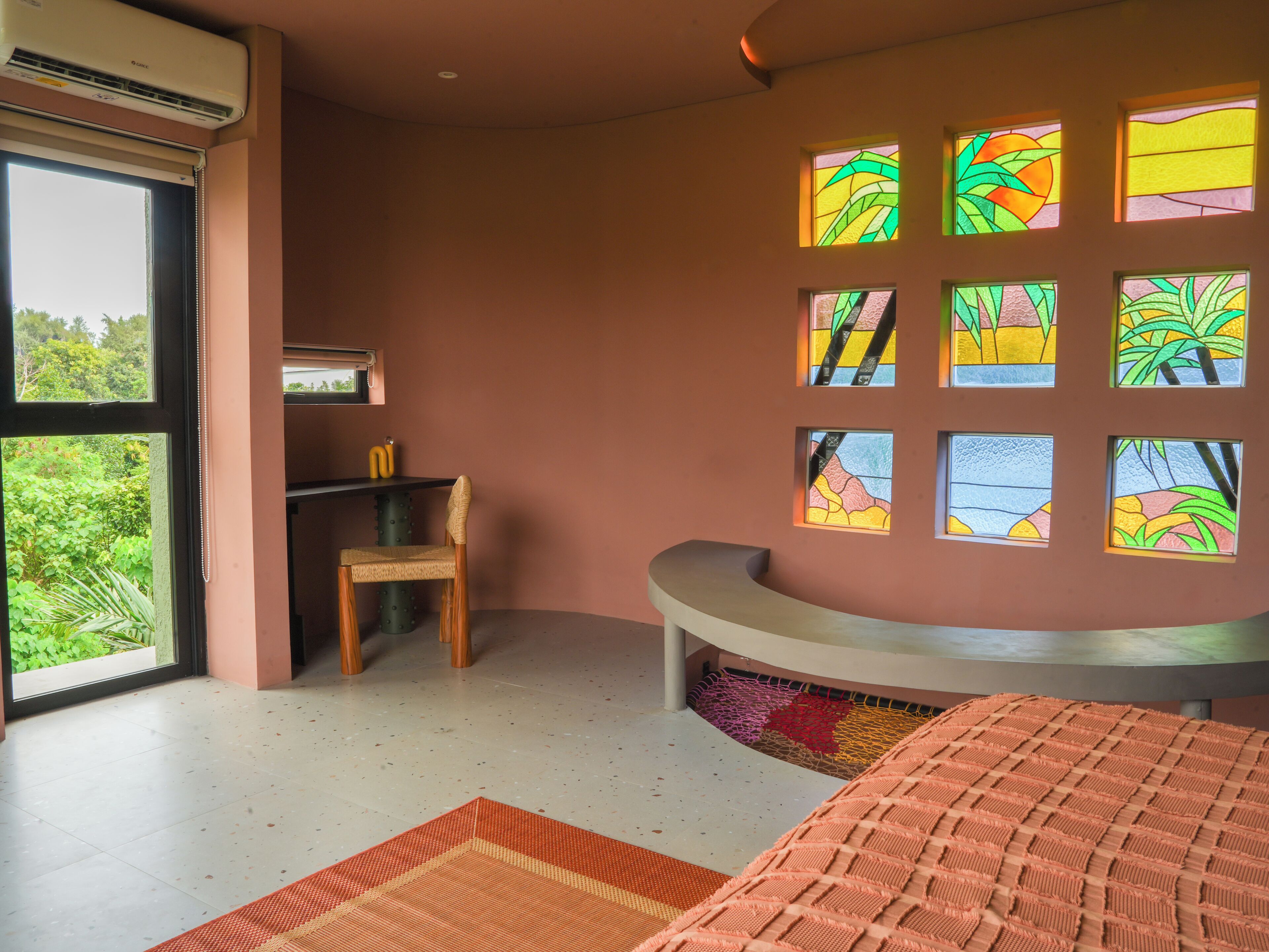 A modern room with a curved bench, a small wooden chair, and a desk with two yellow objects. There are multiple stained glass windows on the wall.