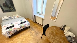 Un dormitorio con cama, piso de madera, ventana, espejo y una mesa de comedor con sillas.