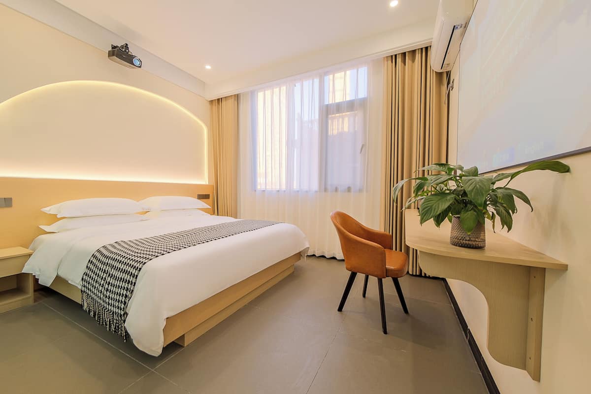 A hotel room with a bed, a desk, a chair, a potted plant, and a projector mounted on the ceiling.