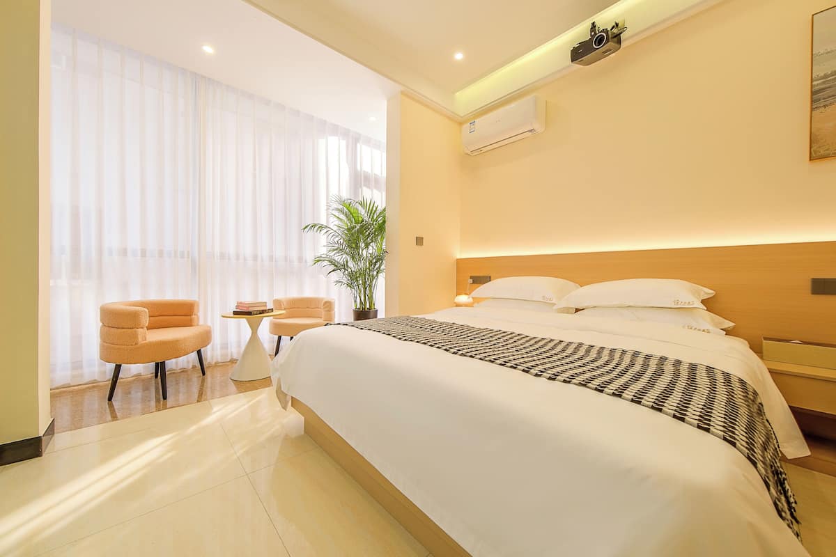 A modern hotel room with a large bed, a small table, a chair, and a potted plant.