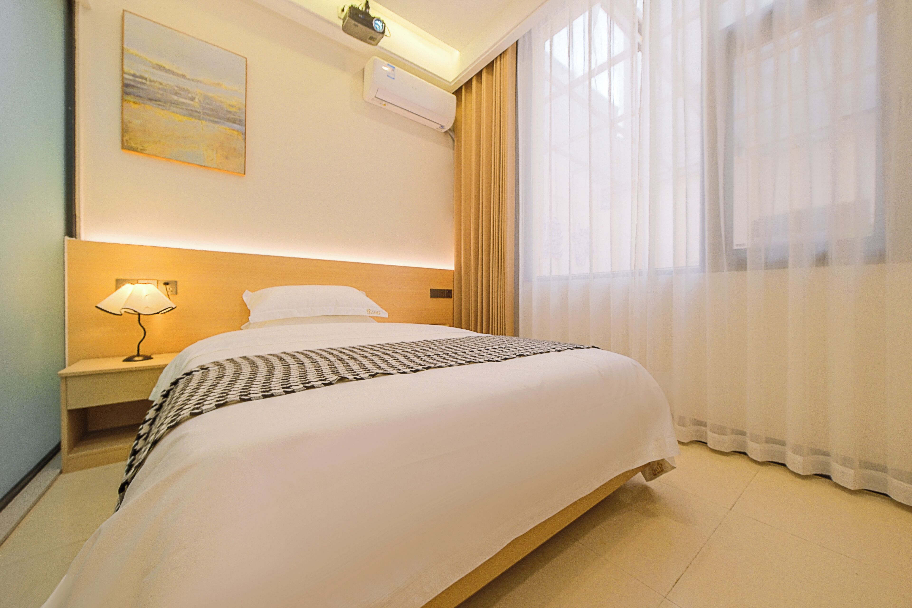 Una habitación de hotel con una cama bien hecha, una lámpara de noche, aire acondicionado de pared y un ventanal con cortinas translúcidas.