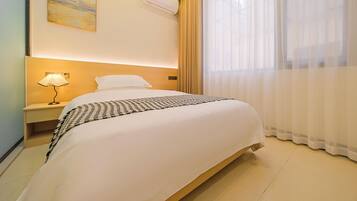 Una habitación de hotel con una cama bien hecha, una lámpara de noche, aire acondicionado de pared y un ventanal con cortinas translúcidas.